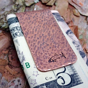 May include: A copper money clip with a hammered finish and a small fish design. The clip is holding a US dollar bill.