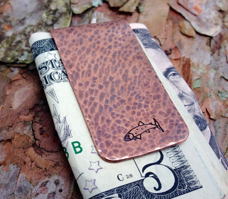 May include: A copper money clip with a hammered finish and a small fish design. The clip is holding a US dollar bill.