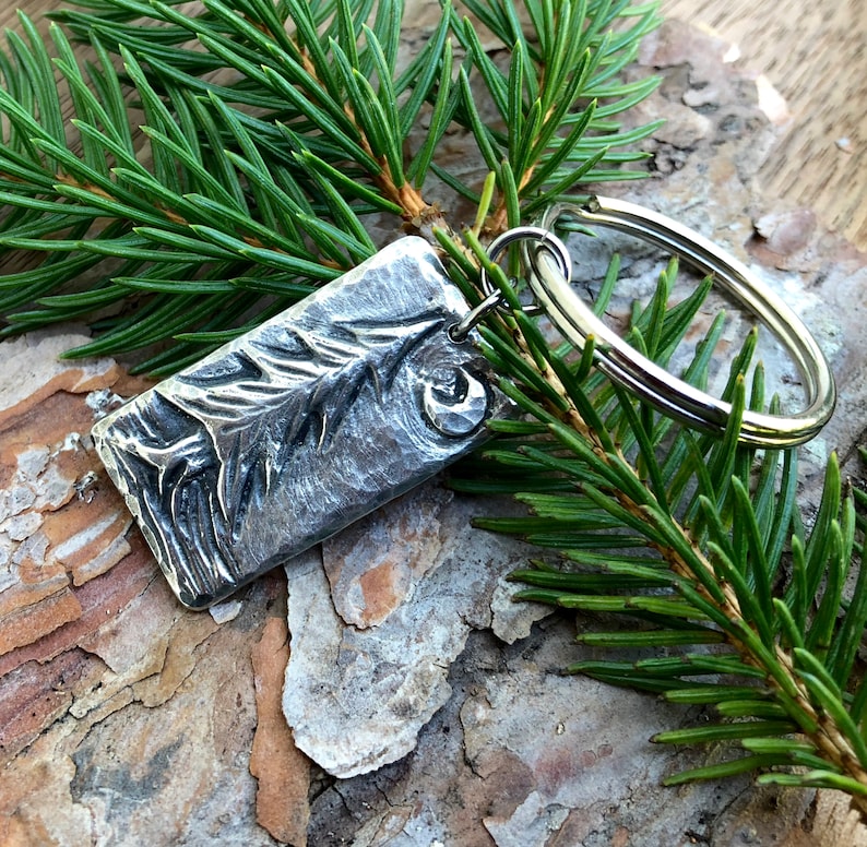 May include: A silver key chain with a rectangular charm featuring a textured design of a pine branch. The charm is attached to a silver key ring.
