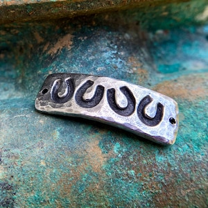 May include: A silver-toned metal charm with three horseshoe shapes, each with a black outline. The charm has a textured surface and is slightly curved.
