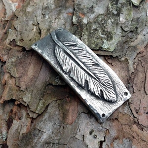 May include: A silver metal pendant with a detailed feather design. The pendant has four small holes around the edge.