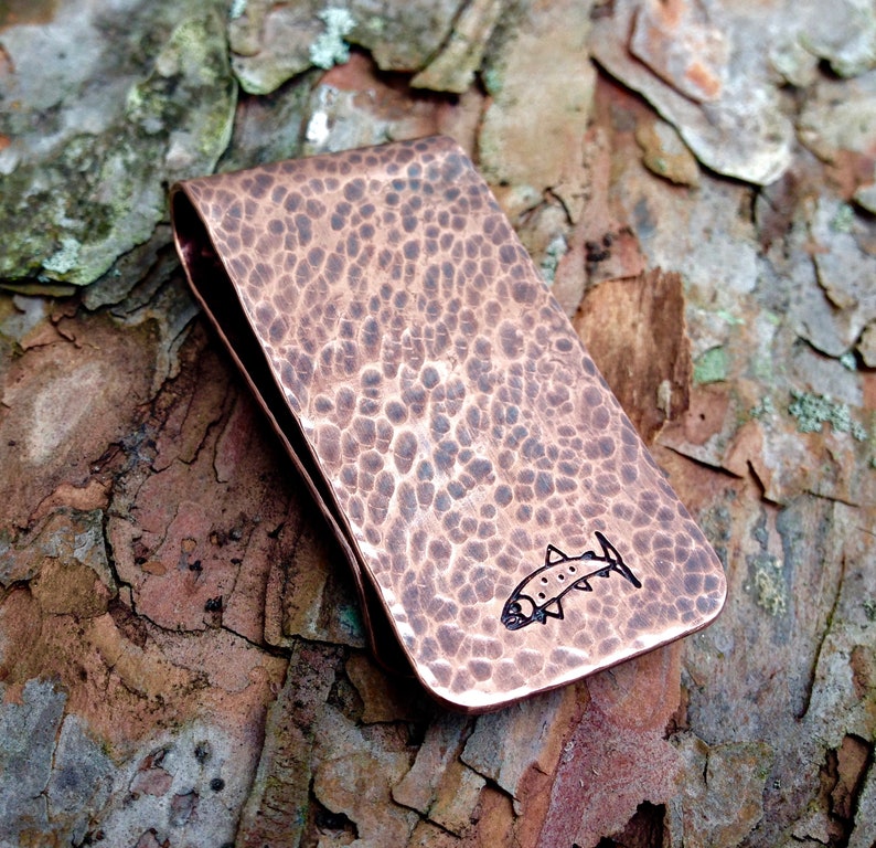 May include: A hammered copper money clip with a fish design. The clip is a dark copper color and has a textured surface.