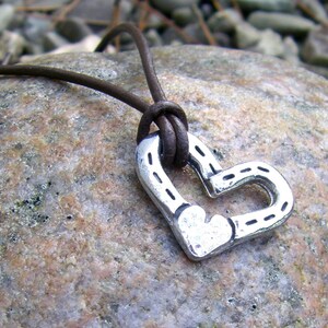 May include: A silver horseshoe heart pendant necklace on a brown leather cord. The pendant is a heart shape made of two horseshoes.
