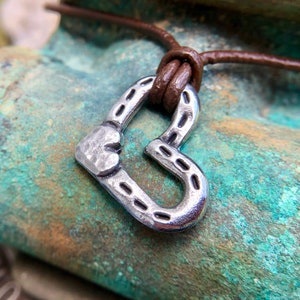 May include: A silver heart-shaped pendant with a textured surface hangs from a brown leather cord. The pendant has a horseshoe design with a small heart at the top.