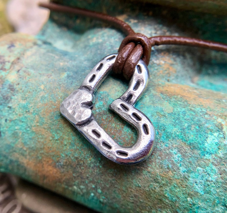 May include: A silver heart-shaped pendant with a textured surface hangs from a brown leather cord. The pendant has a horseshoe design with a small heart at the top.