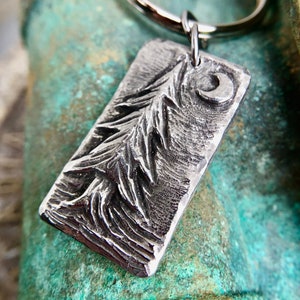 May include: A silver metal keychain with a rectangular tag featuring a detailed embossed design of a pine tree and a crescent moon.