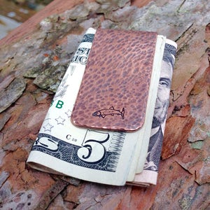 May include: A copper money clip with a hammered finish and a small fish design. The clip is holding a folded five dollar bill.