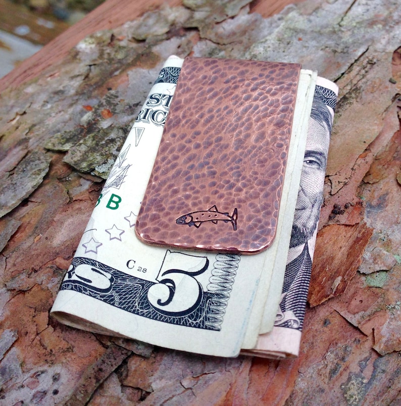 May include: A copper money clip with a hammered finish and a small fish design. The clip is holding a folded five dollar bill.