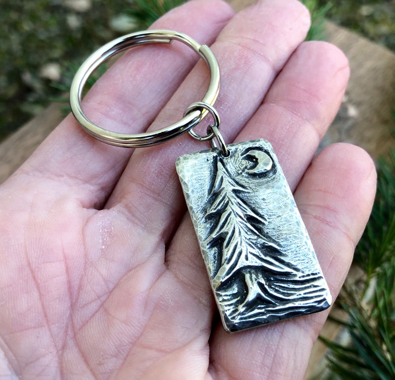 May include: A silver metal keychain with a rectangular charm featuring a detailed tree design and a crescent moon. The charm has a hammered texture.
