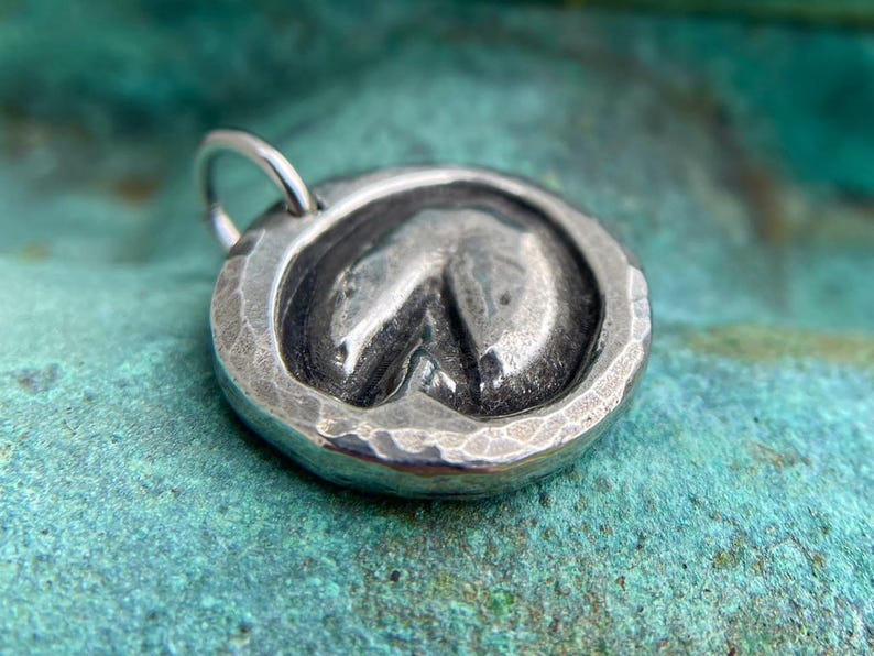 May include: A silver charm in the shape of a horseshoe, with a hammered texture. The charm has a loop for attaching to a necklace or bracelet.