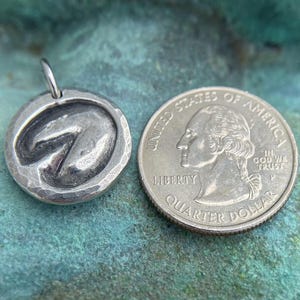 May include: A silver charm in the shape of a horseshoe, next to a US quarter dollar coin for size comparison. The charm has a hammered texture and a slightly raised horseshoe design.