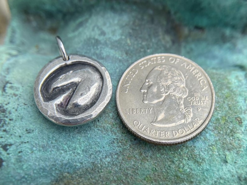 May include: A silver charm in the shape of a horseshoe, next to a US quarter dollar coin for size comparison. The charm has a hammered texture and a slightly raised horseshoe design.
