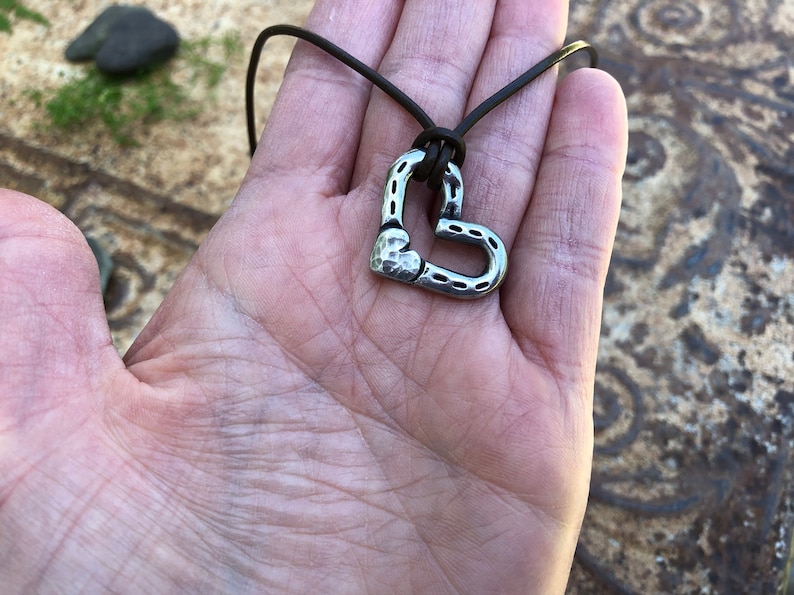 May include: A silver heart-shaped pendant necklace with a hammered texture, hanging from a brown leather cord.