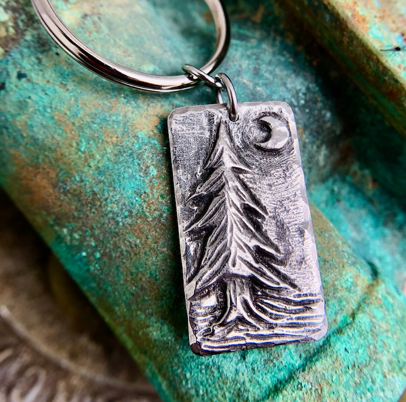 May include: A silver metal keychain with a rectangular charm featuring a detailed embossed design of a pine tree and a crescent moon.