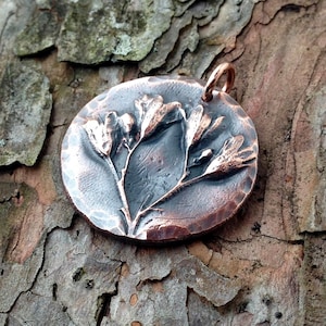May include: A copper pendant with a hammered texture features a raised design of a delicate flower with five petals. The pendant has a loop at the top for attaching a chain.