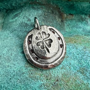 May include: A circular silver-toned metal pendant featuring a raised four-leaf clover design centered within a horseshoe shape. The surface has a textured, hammered finish with dark oxidized detailing in the recessed areas and a small bail at the top.
