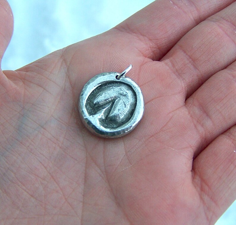 May include: A silver pendant with a textured, circular design featuring a raised, arrow-shaped symbol. The pendant is hanging from a silver chain.