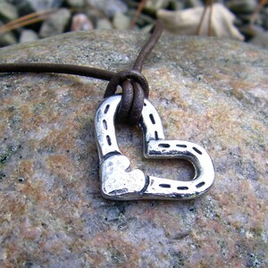 May include: A brown leather cord necklace with a silver heart-shaped pendant. The pendant is made of two horseshoes that are joined together to form a heart shape.