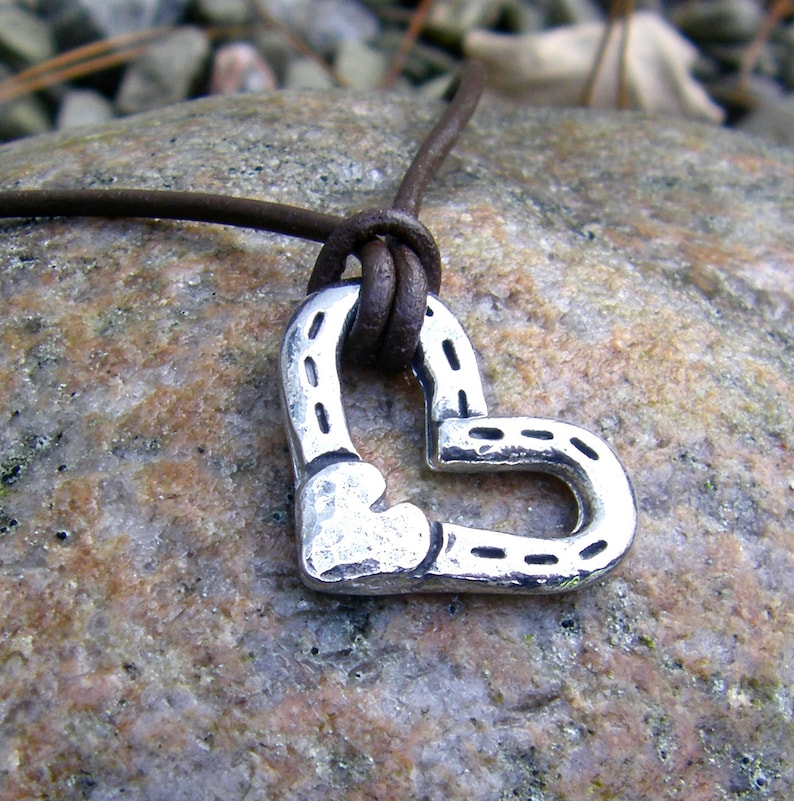 May include: A brown leather cord necklace with a silver heart-shaped pendant. The pendant is made of two horseshoes that are joined together to form a heart shape.