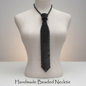Puede incluir: Una corbata de cuentas negras hecha a mano, exhibida en un maniquí. La corbata está construida enteramente con pequeñas cuentas oscuras, creando una apariencia texturizada y sólida. El texto "Handmade Beaded Necktie" está en la parte inferior de la imagen.