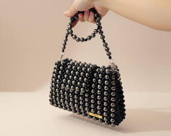 Puede incluir: Un bolso de cuentas de perlas hecho a mano, de forma rectangular, sostenido por una mano. El bolso está hecho de cuentas de color gris oscuro, con un asa de cuentas a juego. El texto "Handmade Pearl Beaded Bag" está en la parte inferior.