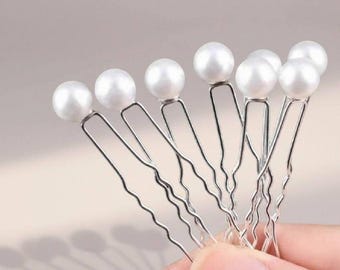 Puede incluir: Un conjunto de horquillas para el cabello con perlas hechas a mano. Cada horquilla presenta una perla blanca y redonda sobre una horquilla de metal plateado ondulado. Las horquillas se sostienen en una mano sobre un fondo suave y neutro. El texto "Handmade Pearl Hair Pins" está en la parte inferior.