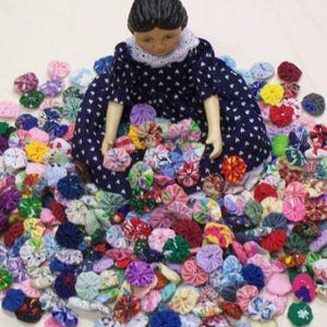May include: A doll in a navy blue dress with white polka dots sits amidst a colorful assortment of fabric yo-yos. The yo-yos are in various colors and patterns, creating a vibrant and textured display.