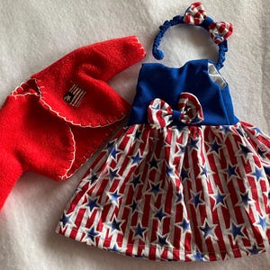 May include: A red fleece bolero jacket with white blanket stitching and a small American flag patch, paired with a sleeveless dress featuring a blue bodice, a red and white striped skirt with blue stars, a matching bow, and a coordinating headband.