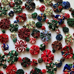 May include: A garland composed of numerous fabric yo-yos in various sizes, featuring festive holiday patterns in red, green, blue, and white. The circular fabric pieces are connected in a continuous string, displaying prints of snowmen, stars, and stripes.