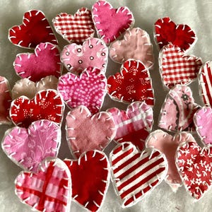 May include: A collection of fabric heart-shaped ornaments in various shades of red and pink. The hearts feature different patterns, including stripes, gingham, and floral designs, with white stitching around the edges. These ornaments are perfect for Valentine's Day or other craft projects.