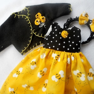 May include: A black felt bolero jacket with yellow blanket stitching and a fabric flower accent, a dress with a black and white polka dot bodice and a yellow skirt featuring a bee pattern, and a matching black and yellow bee print headband.