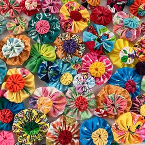 May include: A collection of numerous fabric yo-yos in various sizes, colors, and patterns. Each circular piece is crafted from gathered fabric, featuring a central gathered knot. The assortment includes floral, geometric, and solid-colored textiles.