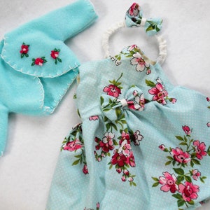 May include: A light blue dress with a floral print and white polka dots. The dress has a bow at the waist and comes with a matching light blue cardigan and a white headband with a floral accent.