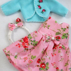 May include: A pink floral dress with a white bow, a light blue cardigan with embroidered flowers, and a white headband with a pink floral bow.