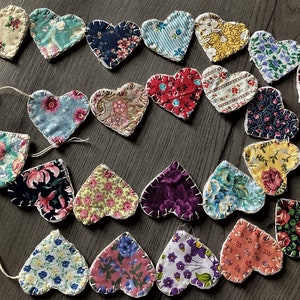 May include: A garland of fabric hearts in various colors and patterns, strung together with white thread. The hearts are made of patchwork fabric and have a rustic, handmade look.