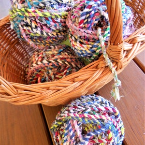 May include: Four balls of colorful yarn made from recycled fabric scraps are in a wicker basket. The yarn is a mix of blues, greens, pinks, reds, and yellows.