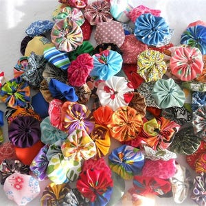 May include: A large collection of fabric yo-yos in a wide variety of colors, patterns, and prints. Each circular fabric piece is gathered in the center to create a ruffled, rosette-like shape, showcasing diverse floral, striped, and geometric designs.