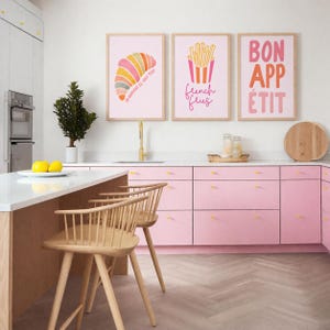 May include: A kitchen scene with three framed prints featuring food-related illustrations and text. The prints hang above a pink cabinet. Two wooden bar stools are in the foreground. The kitchen has a white countertop and a gold faucet.