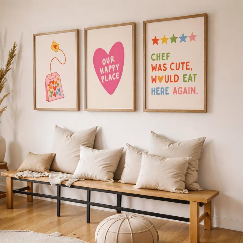 May include: Three framed art prints with whimsical designs hang above a wooden bench with beige pillows. The prints feature a tea bag, a pink heart with text, and colorful text that reads "Chef Was Cute, Would Eat Here Again."