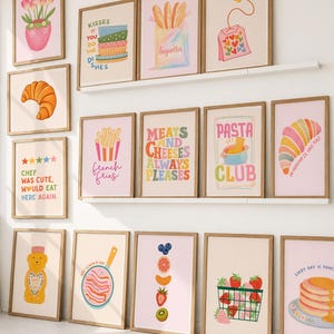 May include: A collection of framed art prints featuring food and kitchen-themed illustrations. Prints include images of croissants, baguettes, strawberries, and text like "French Fries" and "Pasta Club". The frames are a light brown color.