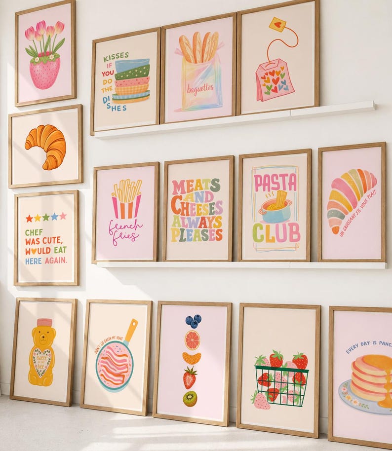 May include: A collection of framed art prints featuring food and kitchen-themed illustrations. Prints include images of croissants, baguettes, strawberries, and text like "French Fries" and "Pasta Club". The frames are a light brown color.