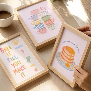 May include: Three framed art prints with light wood frames. One print says "Bake it till you make it" with baking-themed illustrations. Another shows stacked teacups and the text "Tea time?". The third says "Every day is pancake day".