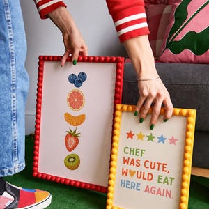 May include: Two framed art prints. One features illustrations of fruit: blueberries, grapefruit, orange slice, strawberry, and kiwi. The other has the text "CHEF WAS CUTE, WOULD EAT HERE AGAIN" with star icons. Both have beaded frames.