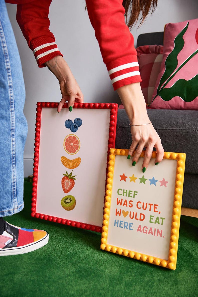 May include: Two framed art prints. One features illustrations of fruit: blueberries, grapefruit, orange slice, strawberry, and kiwi. The other has the text "CHEF WAS CUTE, WOULD EAT HERE AGAIN" with star icons. Both have beaded frames.