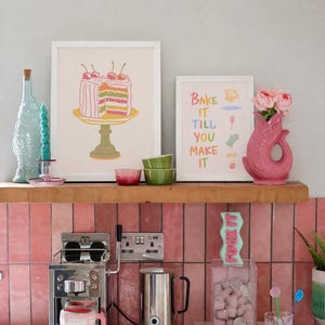May include: A kitchen scene with a pink tiled backsplash and a wooden shelf displaying art prints, a pink vase with flowers, and a coffee maker. The prints read "Bake it till you make it" and a cake. A clear container with marshmallows and a candle that says "FUCK IT" are also visible.