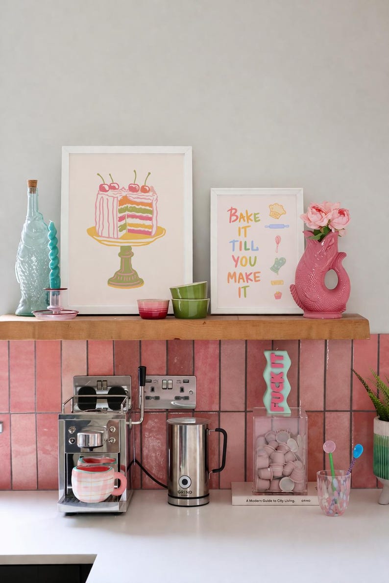 May include: A kitchen scene with a pink tiled backsplash and a wooden shelf displaying art prints, a pink vase with flowers, and a coffee maker. The prints read "Bake it till you make it" and a cake. A clear container with marshmallows and a candle that says "FUCK IT" are also visible.