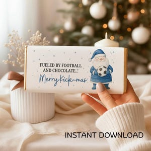 May include: A chocolate bar with a white wrapper featuring a blue Santa Claus holding a football. The text reads "Fueled by Football and Chocolate... Merry Kick-mas." The design includes blue stars and is a festive holiday theme.