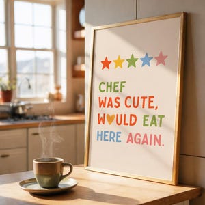 May include: A framed print with the text "CHEF WAS CUTE, WOULD EAT HERE AGAIN" in colorful letters. Above the text are five stars in orange, yellow, green, blue, and pink. A cup of coffee sits on a saucer in the foreground.