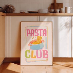May include: A framed poster with the words "PASTA CLUB" in pink, green, and orange. The image features a blue pot with pasta and steam rising from it. The poster is set against a white cabinet.
