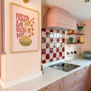 May include: A pink kitchen with a framed poster that says "Pasta La Vista Baby". The kitchen features pink cabinets, a white countertop, and a red and white checkered backsplash. A gold light fixture is mounted above the poster.